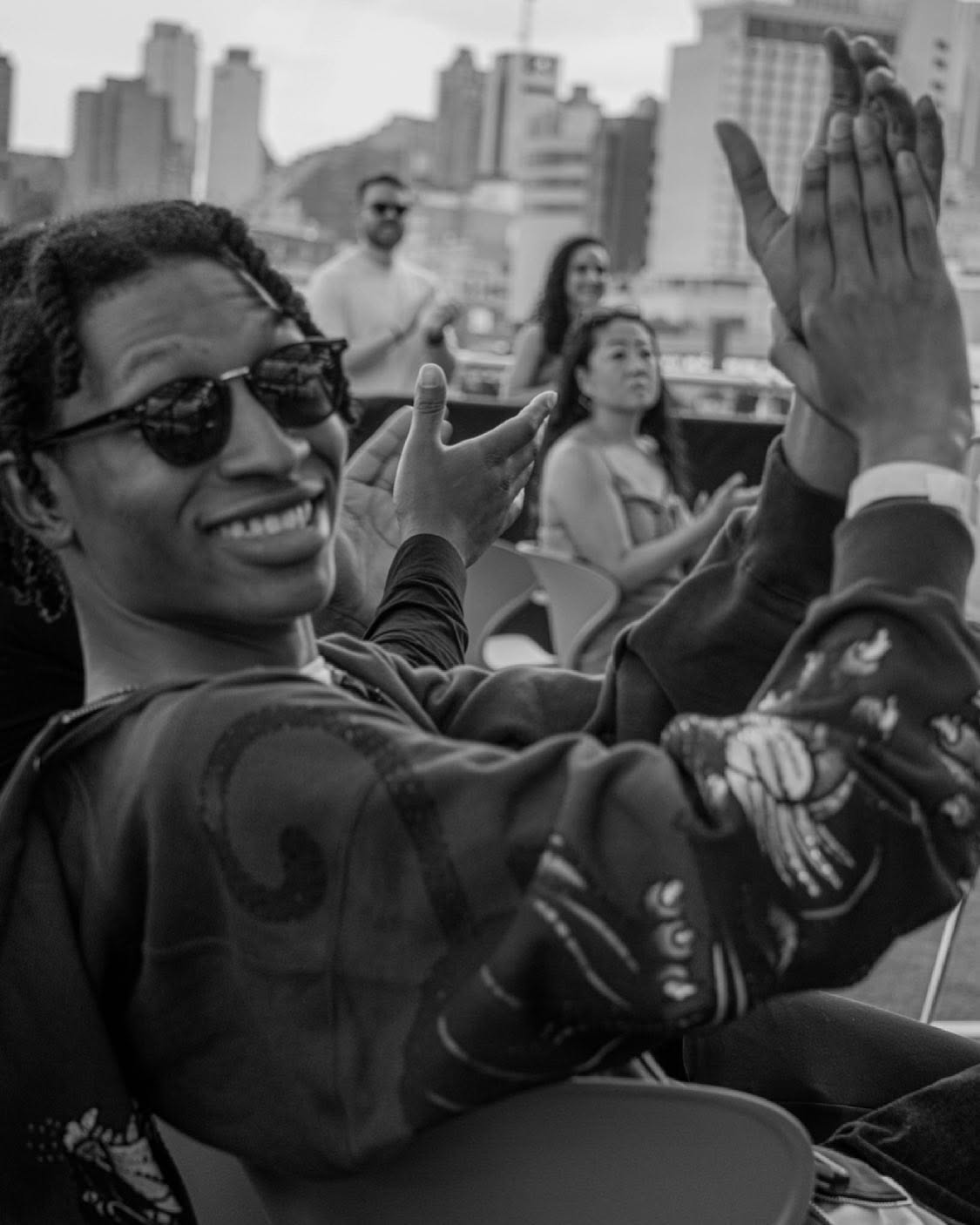 Black-and-white photograph of an outdoor community screening: a young viewer in sunglasses laughs and applauds while neighbors clap behind, framed against a city skyline